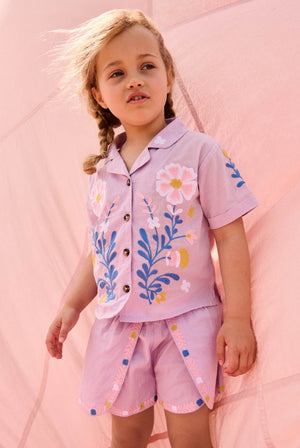A young child with braided hair wears the Louise Misha Girls’ Alova Shirt—a light pink, short-sleeved floral top—along with matching shorts, standing in front of a pale pink fabric backdrop.