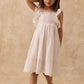 A young child with straight brown hair wears the Louise Misha Girls’ Anaya Dress—a white cotton dress with a smocked yoke and delicate eyelet embroidery—paired with gold sandals, standing against a beige textured background.