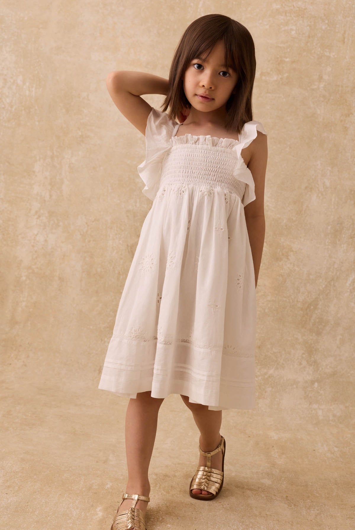A young child with straight brown hair wears the Louise Misha Girls’ Anaya Dress—a white cotton dress with a smocked yoke and delicate eyelet embroidery—paired with gold sandals, standing against a beige textured background.