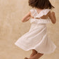 A girl wearing the Louise Misha Girls’ Anaya Dress—crafted in white cotton with delicate eyelet embroidery and a smocked yoke—twirls with her back to the camera against a beige textured background, her sandals softly grounded on the floor.