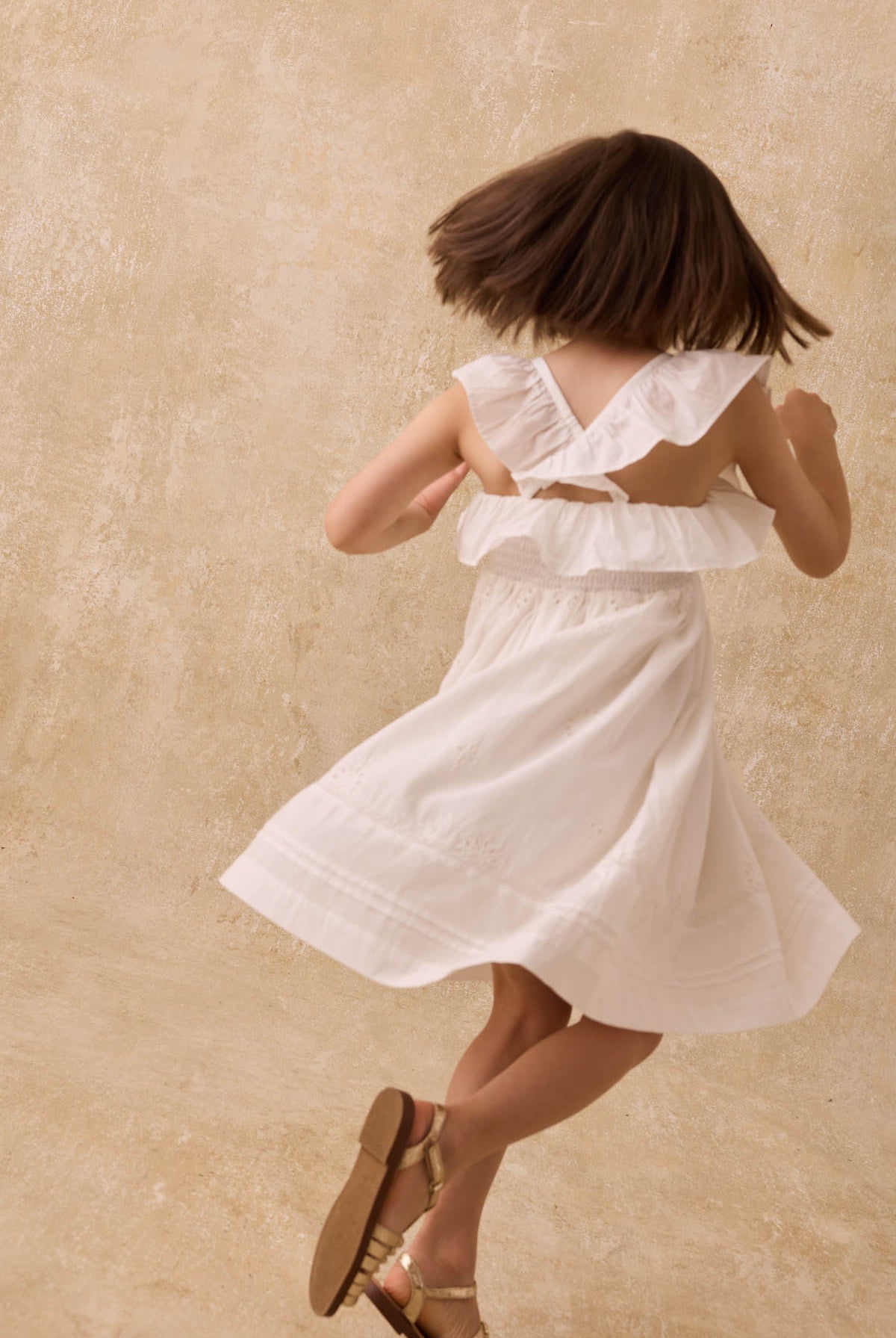 A girl wearing the Louise Misha Girls’ Anaya Dress—crafted in white cotton with delicate eyelet embroidery and a smocked yoke—twirls with her back to the camera against a beige textured background, her sandals softly grounded on the floor.