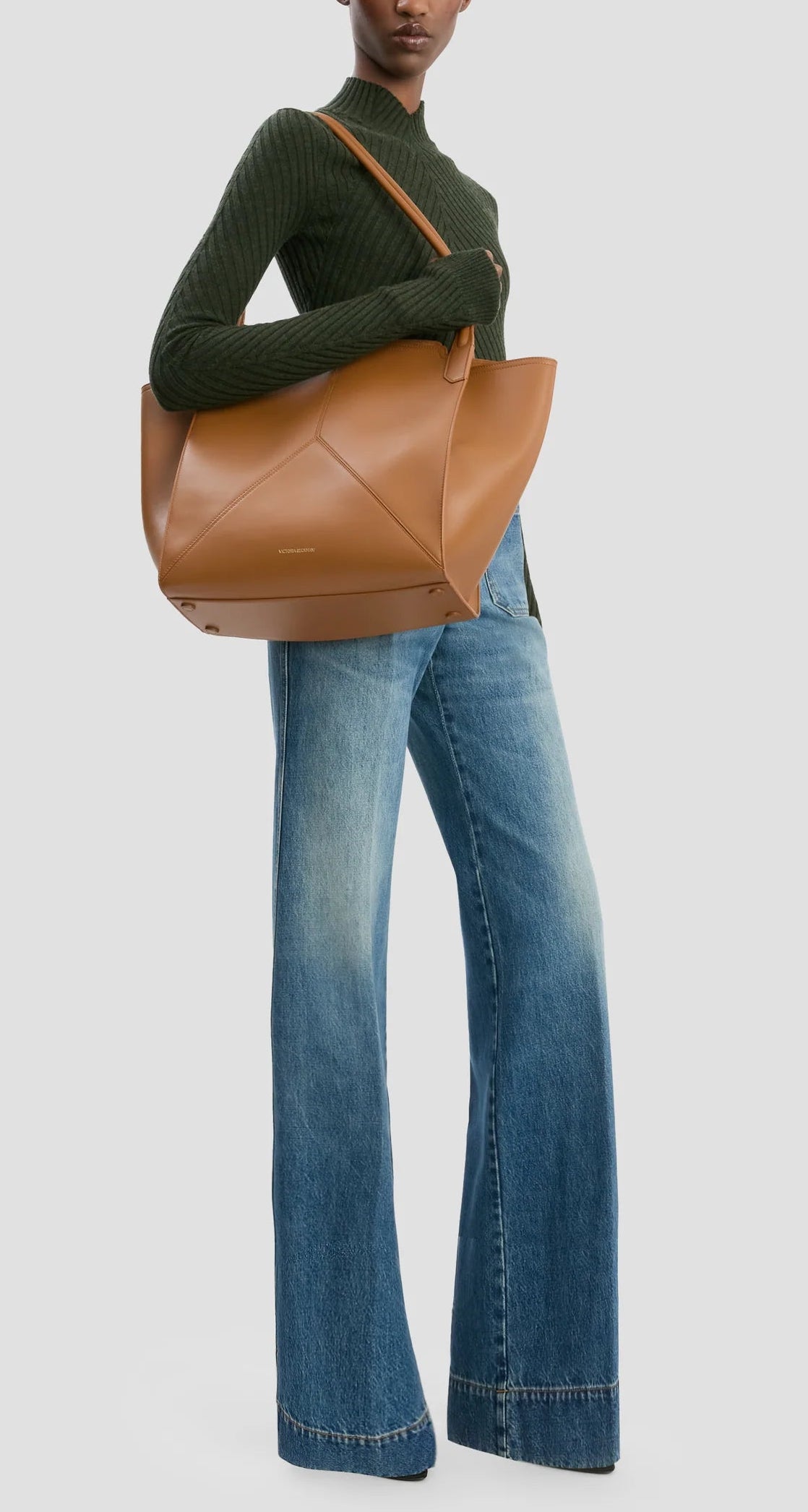 A person in a green ribbed sweater and wide-leg blue jeans carries the Victoria Beckham Small Victoria Tote in tan leather over their shoulder.