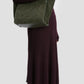 A person in a long-sleeved, ribbed purple dress and brown heels carries the Victoria Beckham Small Victoria Suede Tote in Dark Olive on their shoulder.