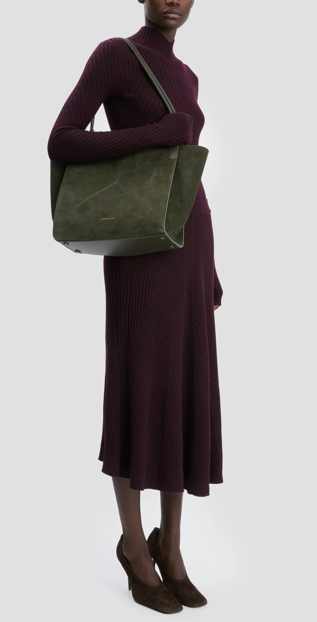 A person in a long-sleeved, ribbed purple dress and brown heels carries the Victoria Beckham Small Victoria Suede Tote in Dark Olive on their shoulder.