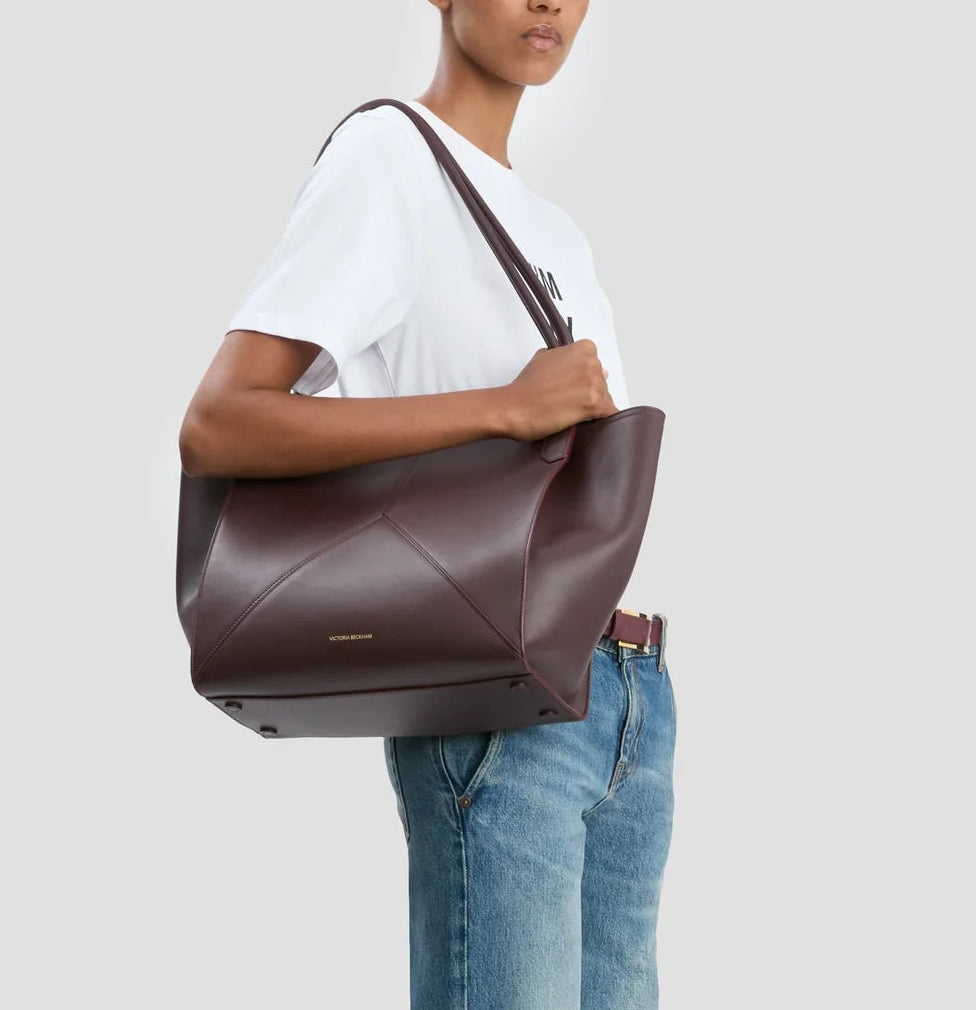 A person in a white t-shirt and blue jeans carries the Victoria Beckham Victoria Tote, a large dark brown handbag lined with suede, on their shoulder.