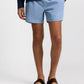 A model's lower body is shown wearing Onia Charles 5" Swim Trunk in light blue, paired with a dark long-sleeve top and dark brown sandals, standing against a plain white background.
