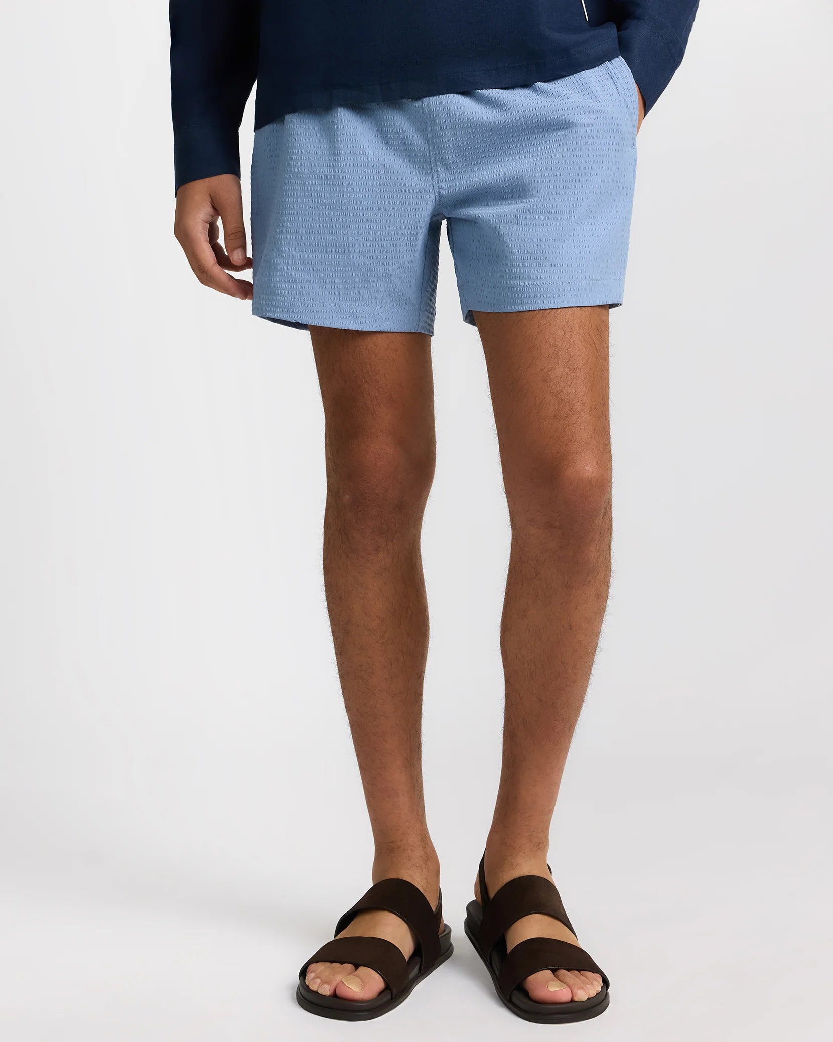 A model's lower body is shown wearing Onia Charles 5" Swim Trunk in light blue, paired with a dark long-sleeve top and dark brown sandals, standing against a plain white background.