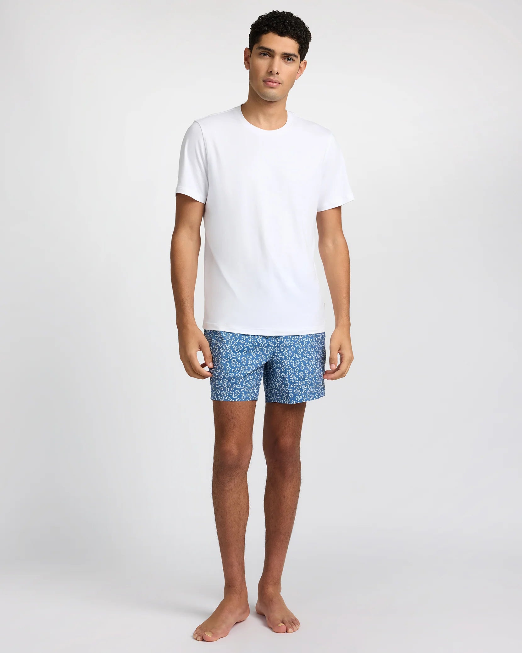 A man stands barefoot against a plain background, wearing a white T-shirt and Onia’s Charles 5" Swim Trunk in blue, crafted from quick-dry fabric.