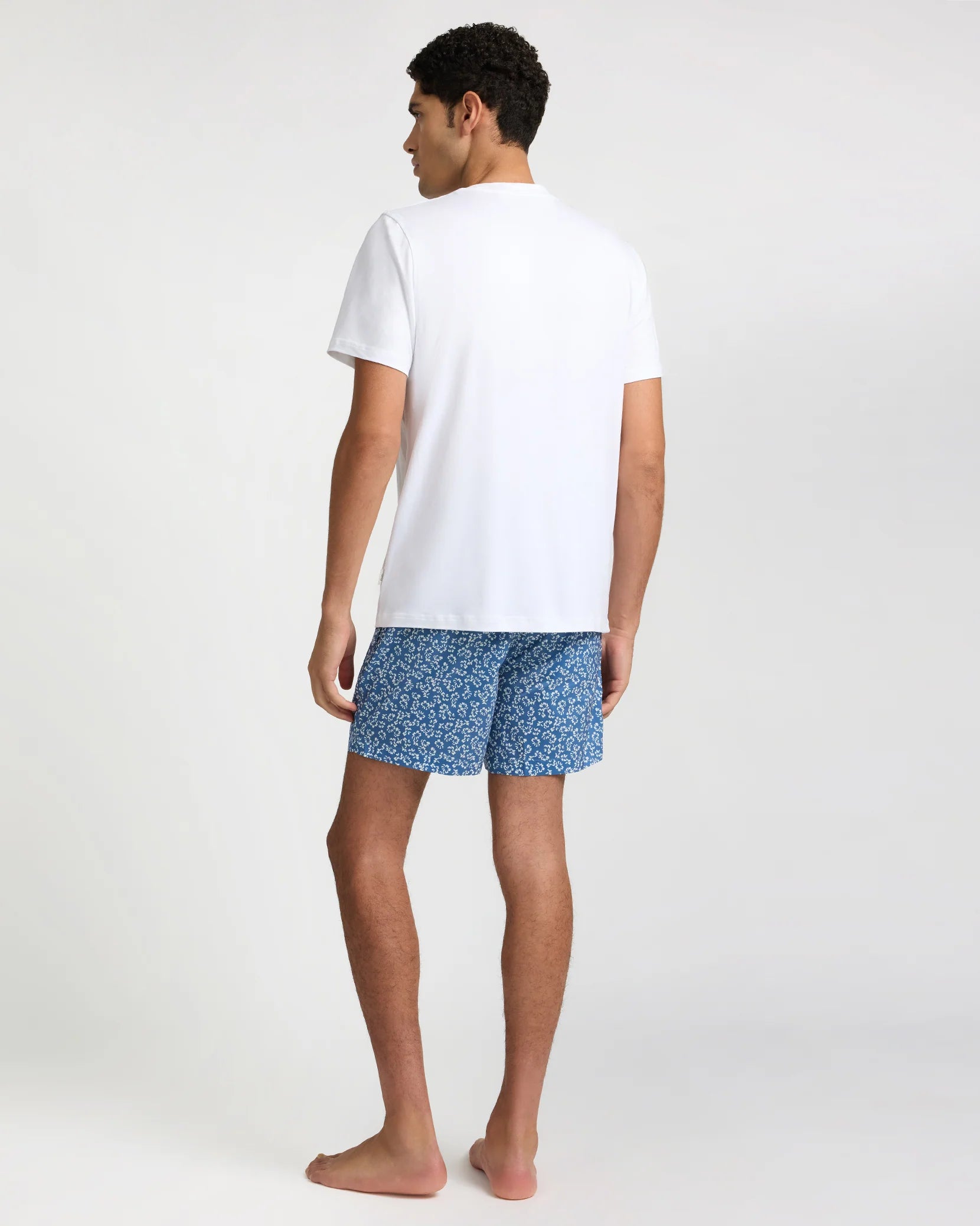 A man stands barefoot facing away, wearing a plain white T-shirt and Onia’s Charles 5" Swim Trunk in blue with classic tailoring, set against a plain light background.