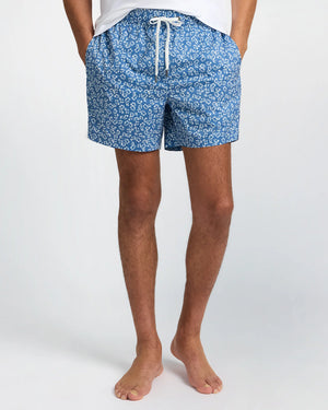 A person wearing Onia Charles 5" Swim Trunk in blue with a white floral pattern, standing barefoot against a plain background.