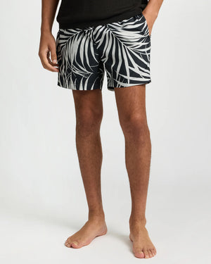 A person stands barefoot in Onia's Charles 5" Swim Trunk, featuring a bold palm leaf pattern and classic tailoring, paired with a black shirt. The photo is cropped above the waist.