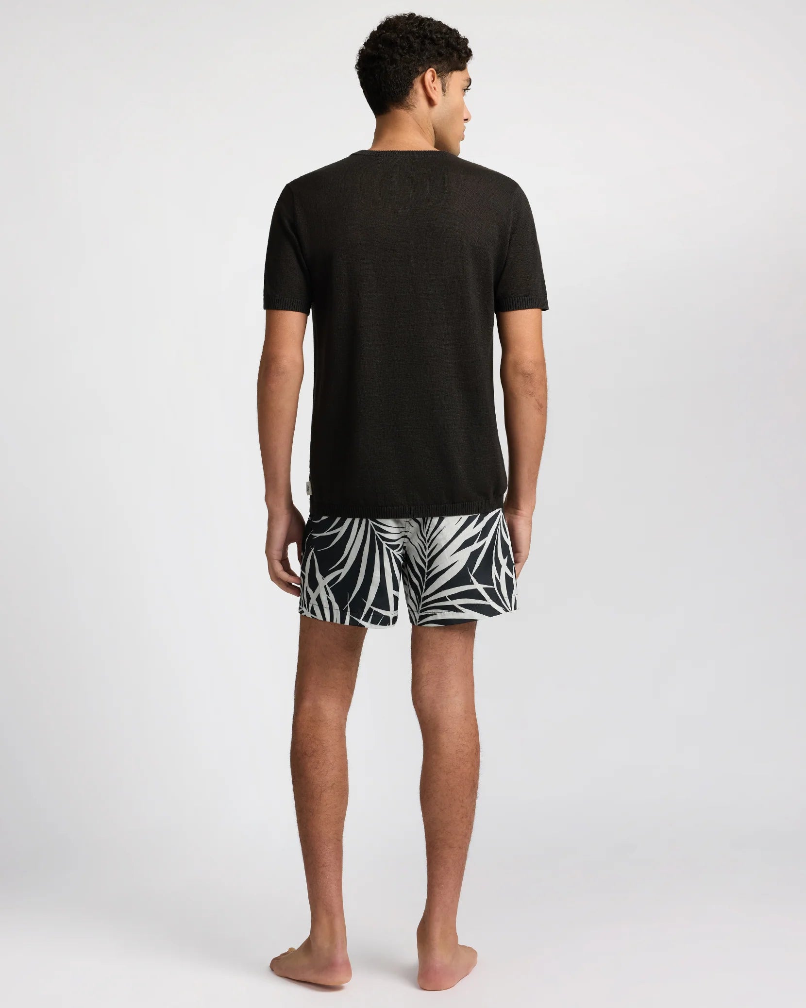 A man stands barefoot facing away, wearing a black short-sleeve shirt and Onia’s Charles 5" Swim Trunk in a black-and-white pattern with classic tailoring, set against a plain light background.