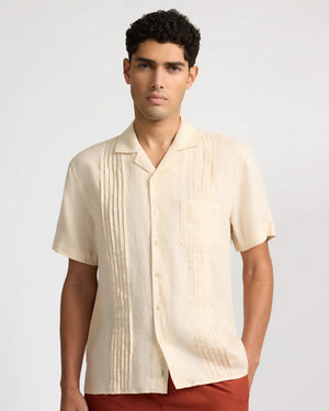 A man wearing Onia's Linen Pintuck Camp Shirt in cream, featuring vertical pintuck pleats and a front pocket, pairs it with reddish-brown pants against a plain background.