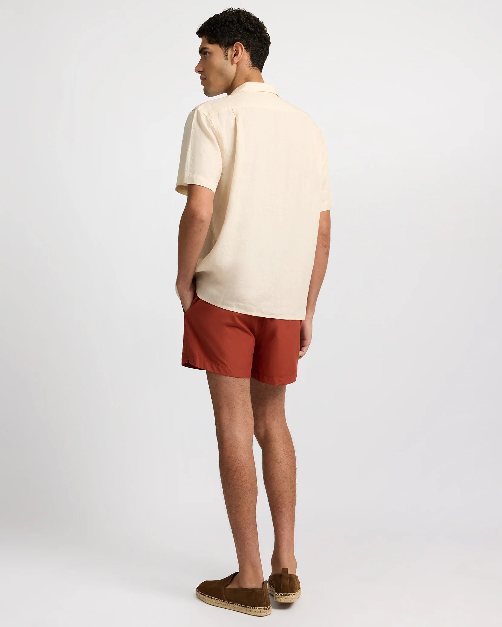 A man faces away, wearing the Onia Linen Pintuck Camp Shirt with pintuck pleats, paired with rust shorts and brown loafers against a plain light background.