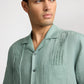 A person in the Onia Linen Pintuck Camp Shirt—a light green pure linen shirt with vertical pintuck pleats and a chest pocket—looks off to the side against a plain background.