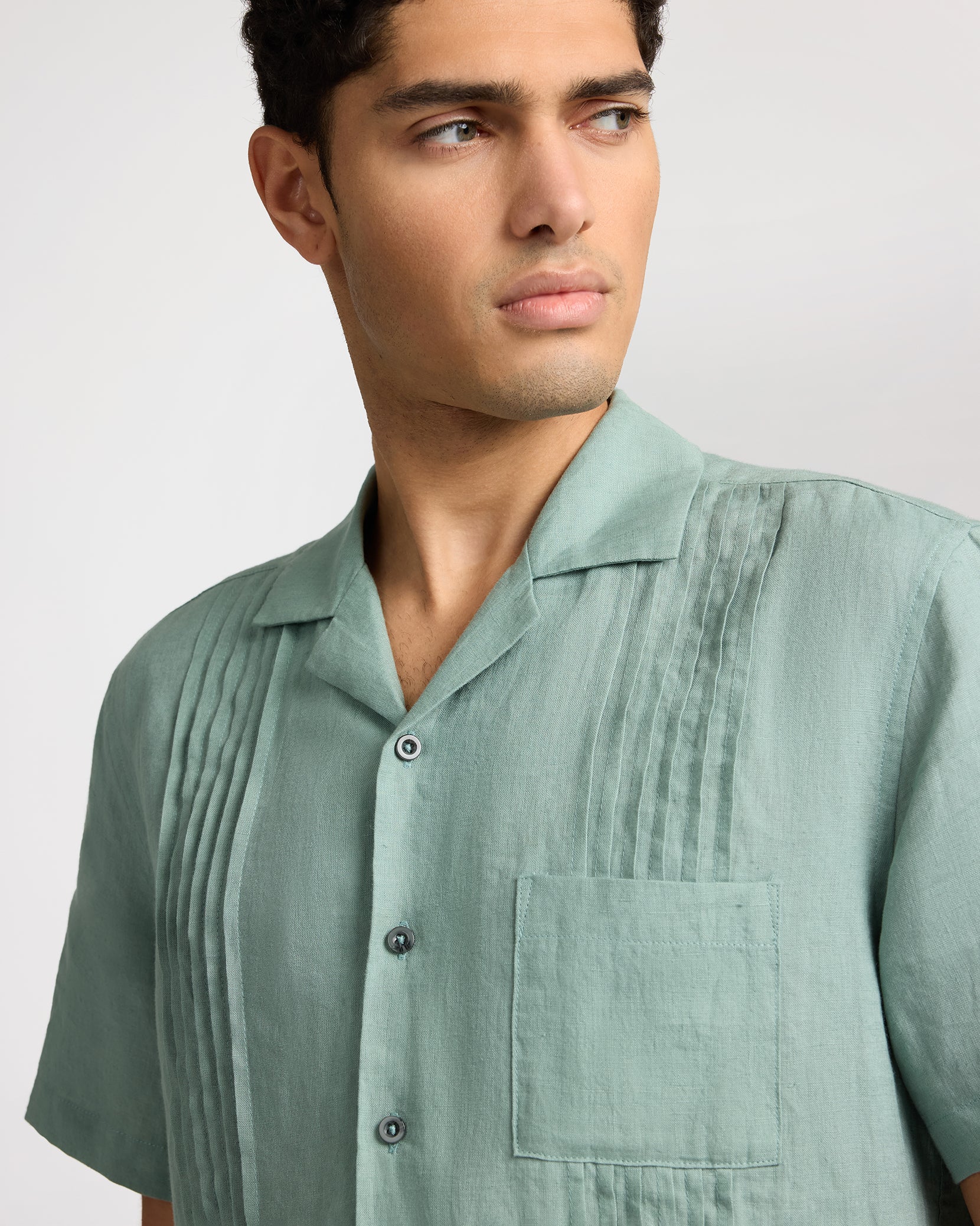 A person in the Onia Linen Pintuck Camp Shirt—a light green pure linen shirt with vertical pintuck pleats and a chest pocket—looks off to the side against a plain background.