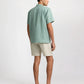 A man stands with his back to the camera, wearing an Onia Linen Pintuck Camp Shirt in light green, paired with beige shorts and brown slip-on shoes, set against a plain background.
