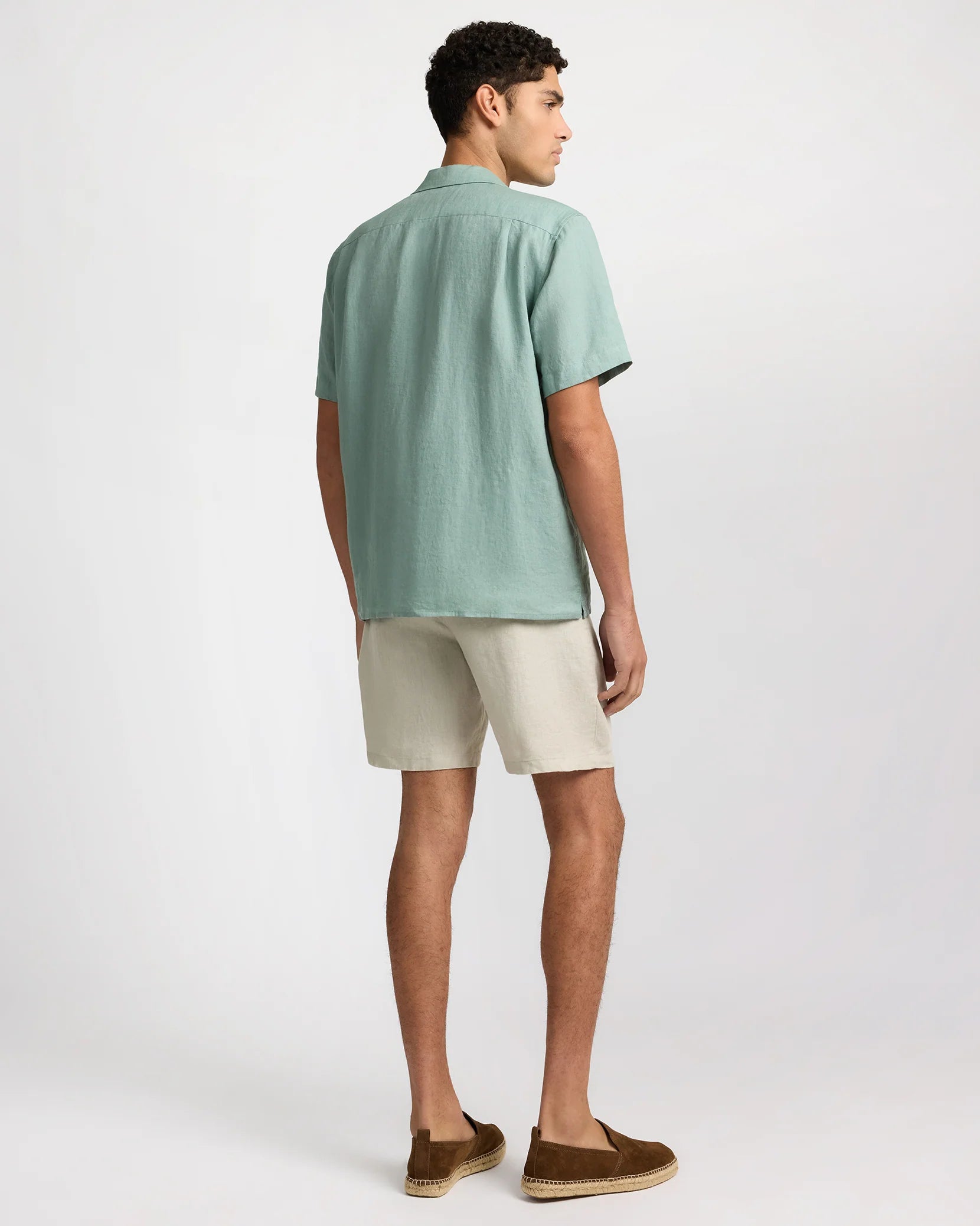 A man stands with his back to the camera, wearing an Onia Linen Pintuck Camp Shirt in light green, paired with beige shorts and brown slip-on shoes, set against a plain background.