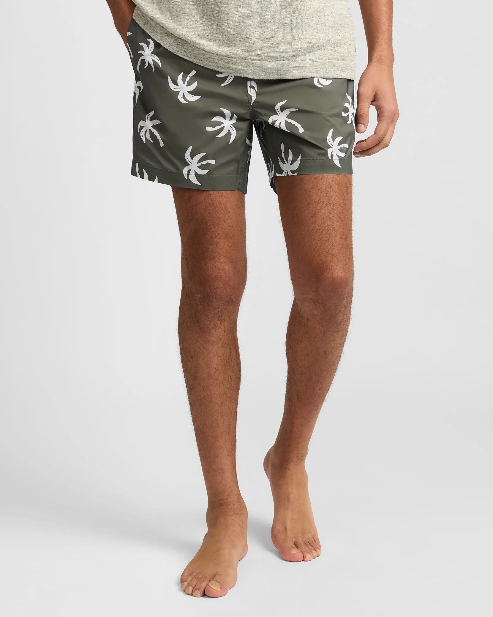 A person stands barefoot against a plain background wearing Onia’s Charles 5" Swim Trunk in olive green with a white palm tree print, made from quick-dry fabric.