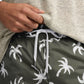 Someone adjusts the drawstring on Onia's Charles 5" Swim Trunk in green with white palm tree prints, made from quick-dry fabric, paired with a textured beige shirt.