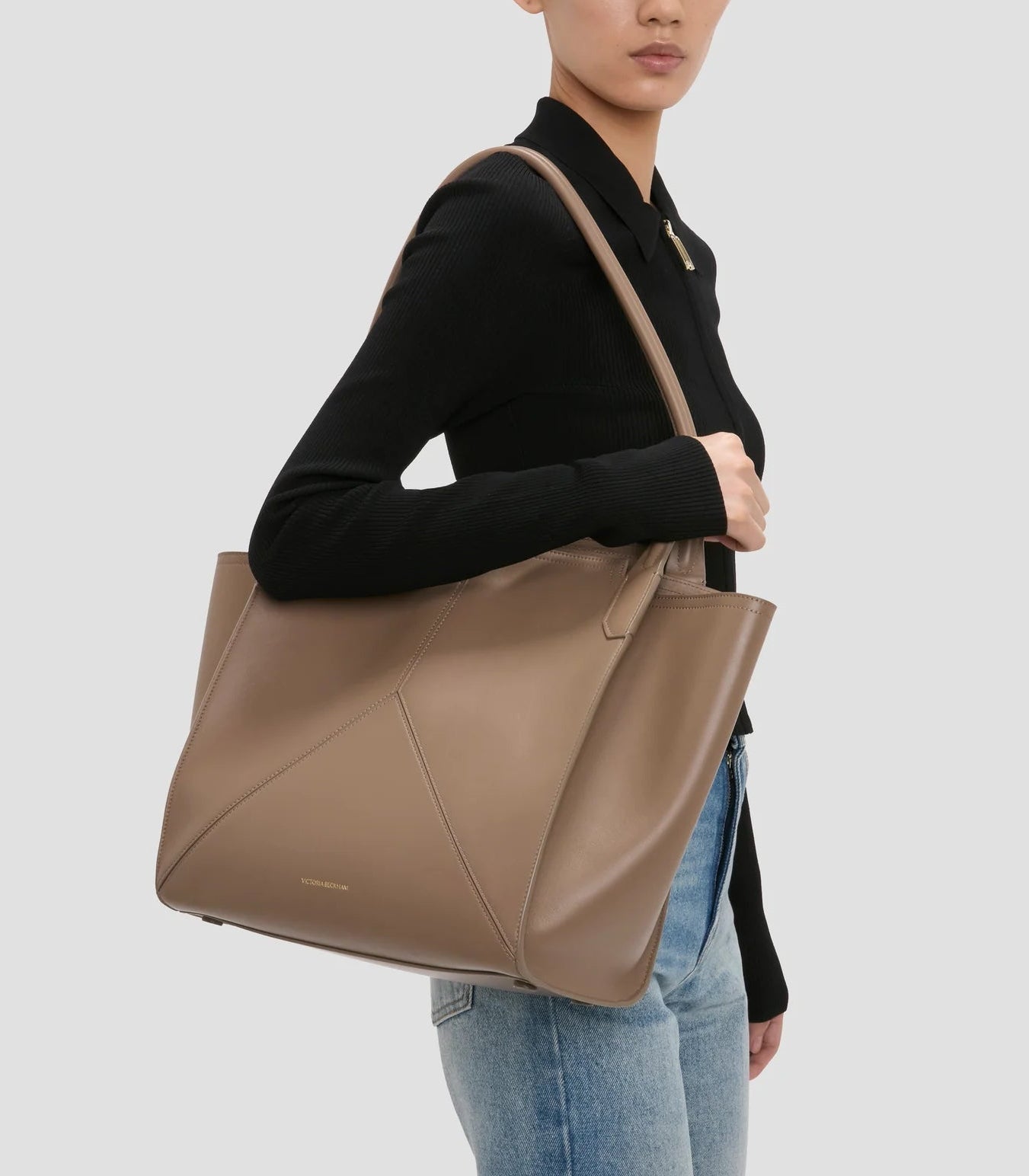 A person in a black long-sleeve top and jeans carries the Victoria Beckham Victoria Tote, a large taupe suede-lined handbag, over their shoulder.