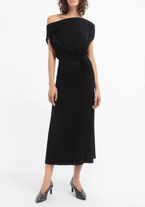 A woman wears the CO Draped Off Shoulder Jersey Dress in black viscose jersey with a knotted waist, styled with black polka dot pointed-toe heels.