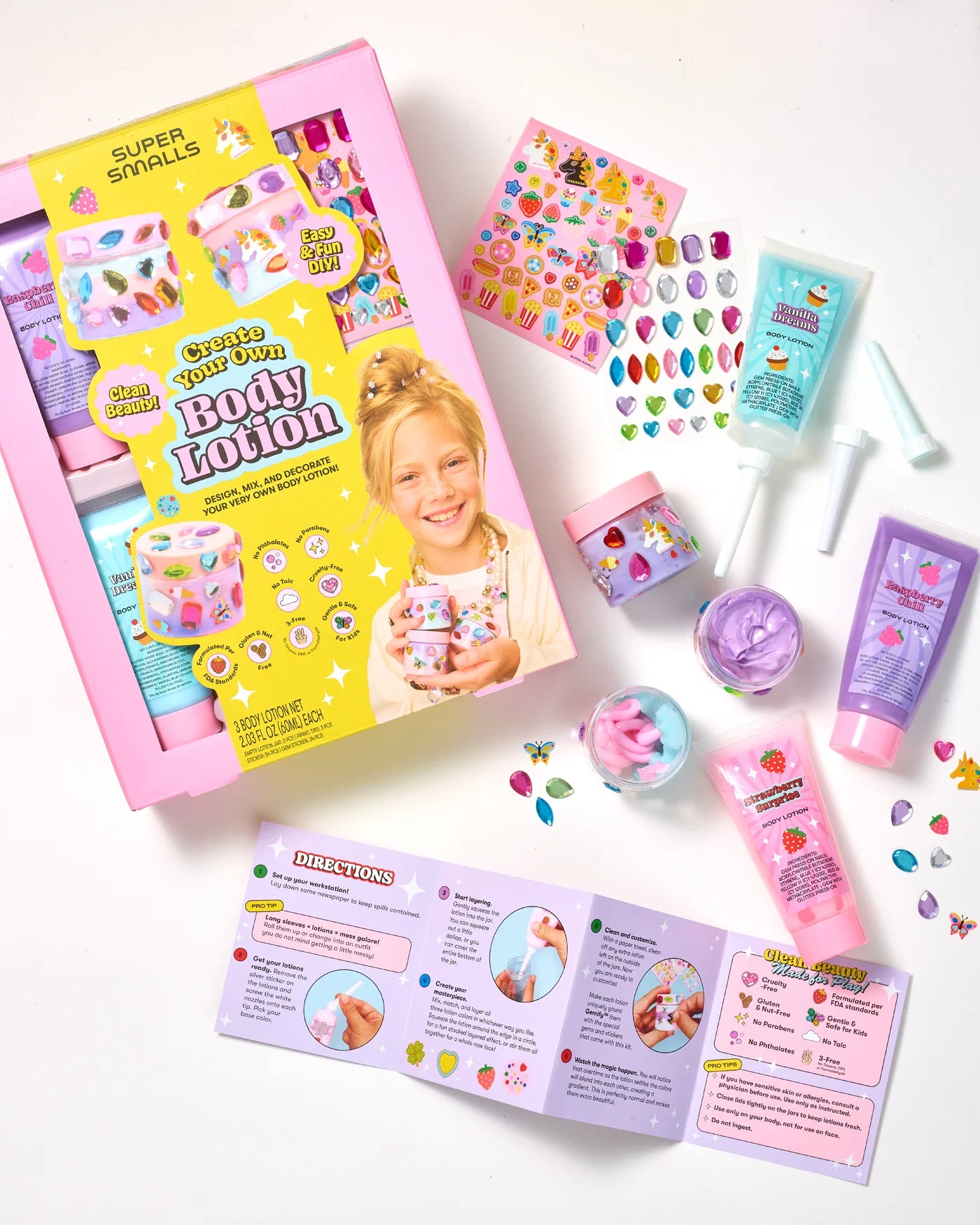 The Super Smalls Create Your Own Body Lotion kit is displayed with lotion tubes, jars, stickers, mixing tools, and an instruction booklet laid out on a white surface. This DIY kit allows kids to safely mix and decorate their own body lotions for a fun and educational beauty activity.