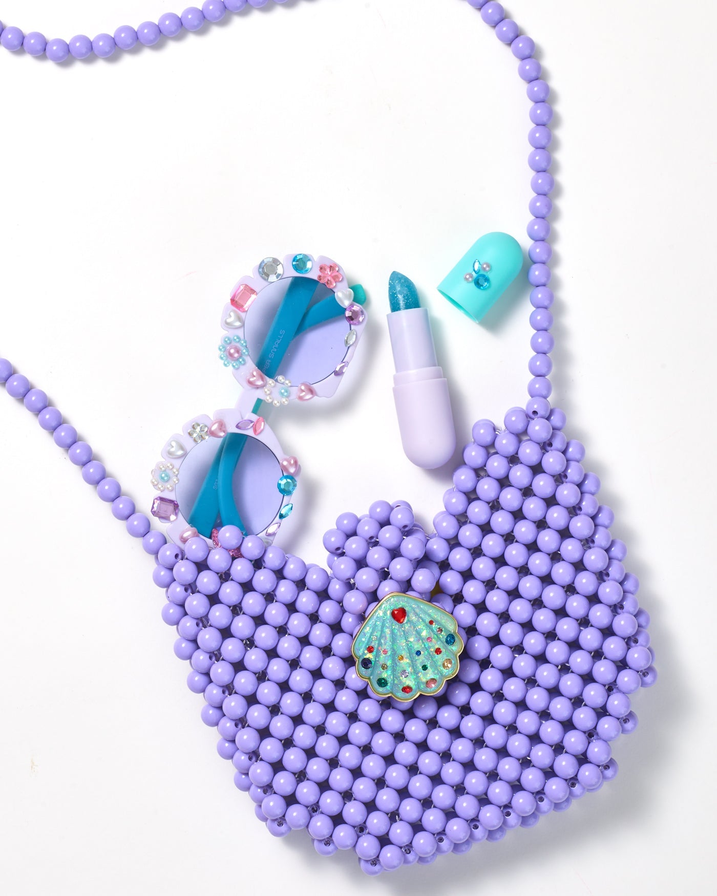 The Super Smalls Mermaid Dreams Handbag, paired with glittery blue lipstick and gem-studded round sunglasses, is stylishly displayed on a white background.