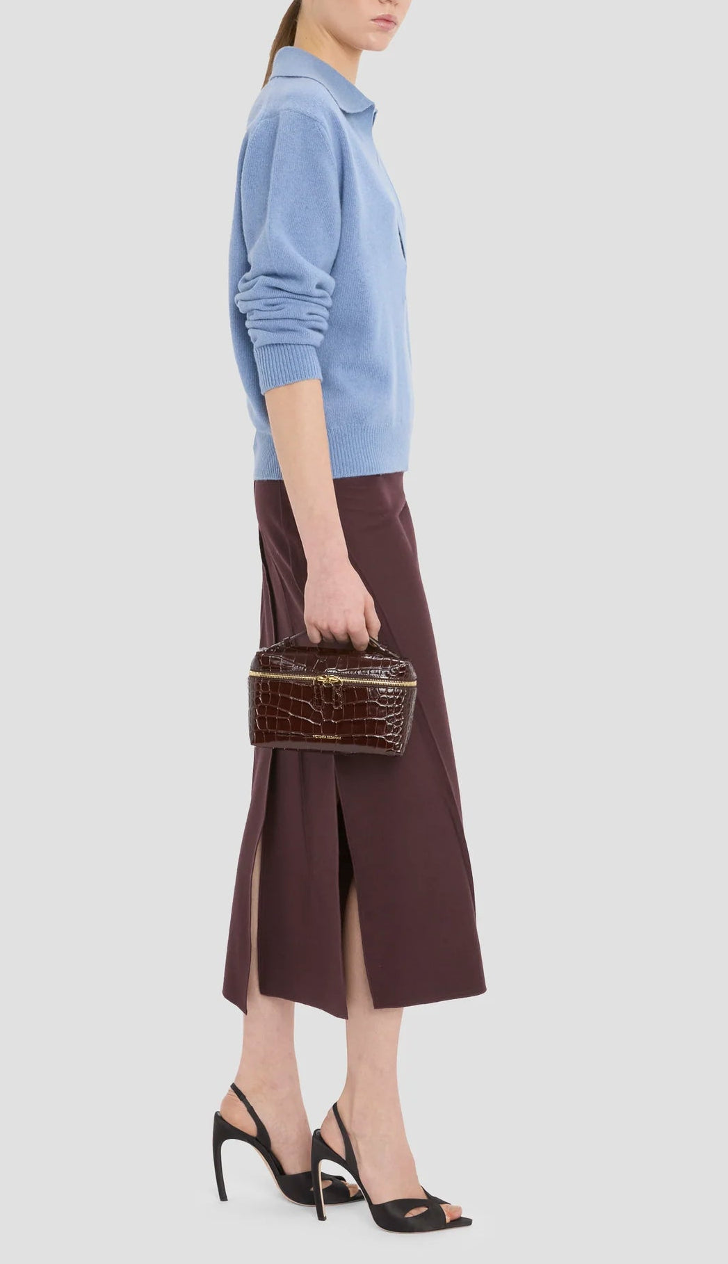 Woman in profile wears a light blue sweater, dark midi skirt with side slits, black heeled sandals, and carries the Victoria Beckham Vanity Croc Crossbody handbag.
