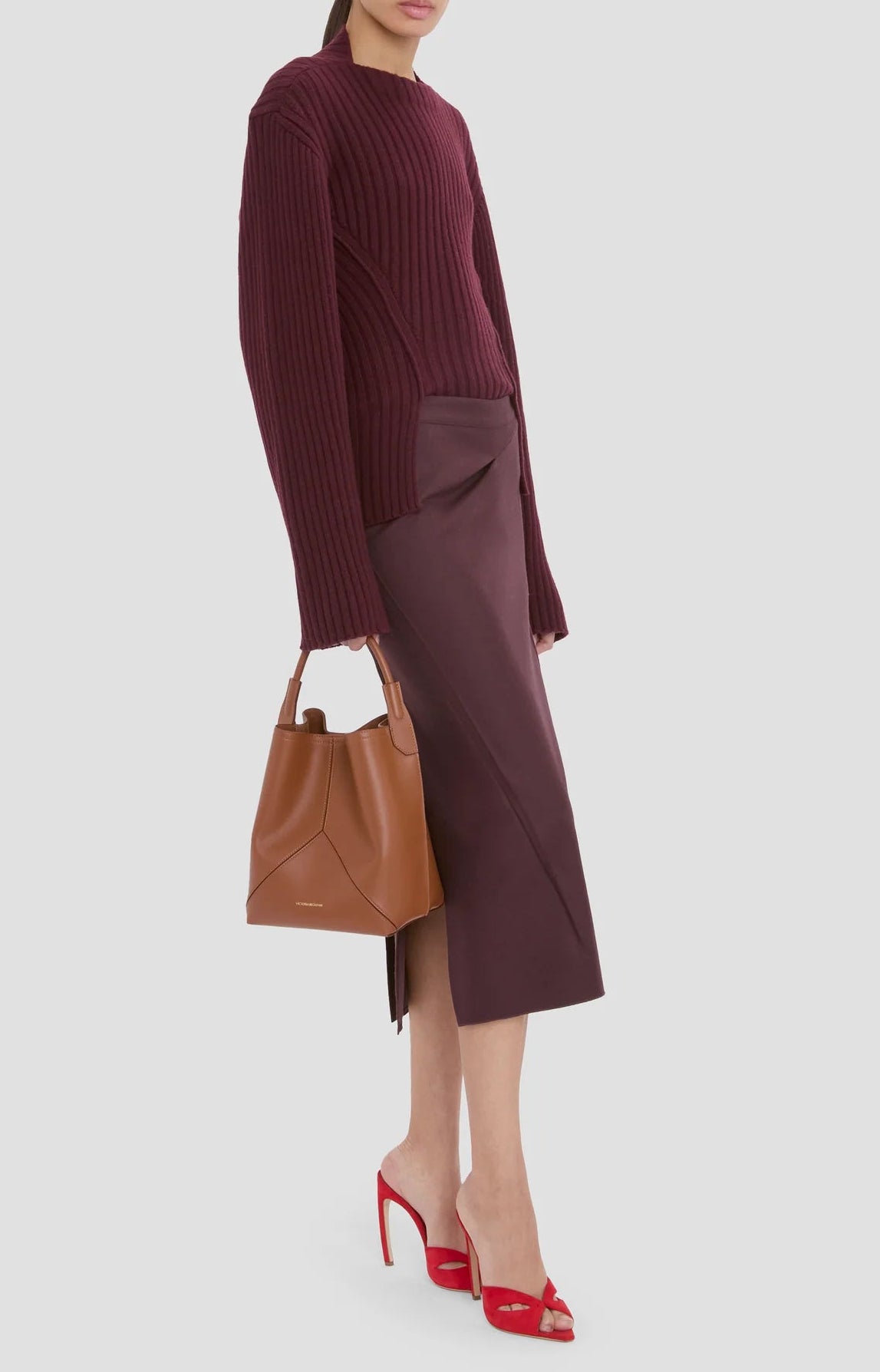 A person in a burgundy ribbed sweater, dark purple midi skirt, red open-toe heels, and carrying the Victoria Beckham Victoria Bucket bag in Dark Tan nappa leather completes this modern look.