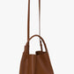 The Victoria Beckham Victoria Bucket in dark tan nappa leather features dual handles, a long shoulder strap, clean lines, and minimal gold branding at the base for a modern look.