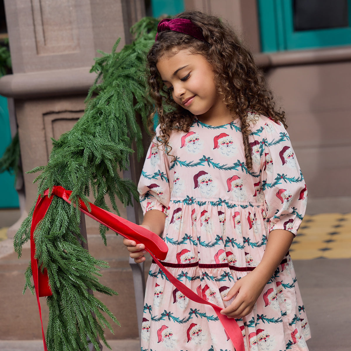 A child wearing a burgundy velour Girls’ Holly Headband and a light pink dress with a Santa Claus print, holding a red ribbon on a garland.