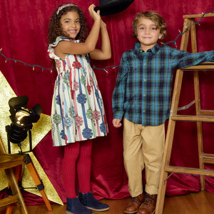 A girl in a floral dress and a boy in a plaid shirt and Pink Chicken Boys’ Jackson Pant standing in front of a red curtain.