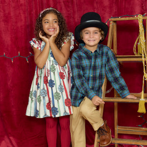 A girl in a patterned dress and a boy in a blue tartan Boys’ Jack Shirt by Pink Chicken and a top hat stand in front of a red curtain and a wooden ladder.