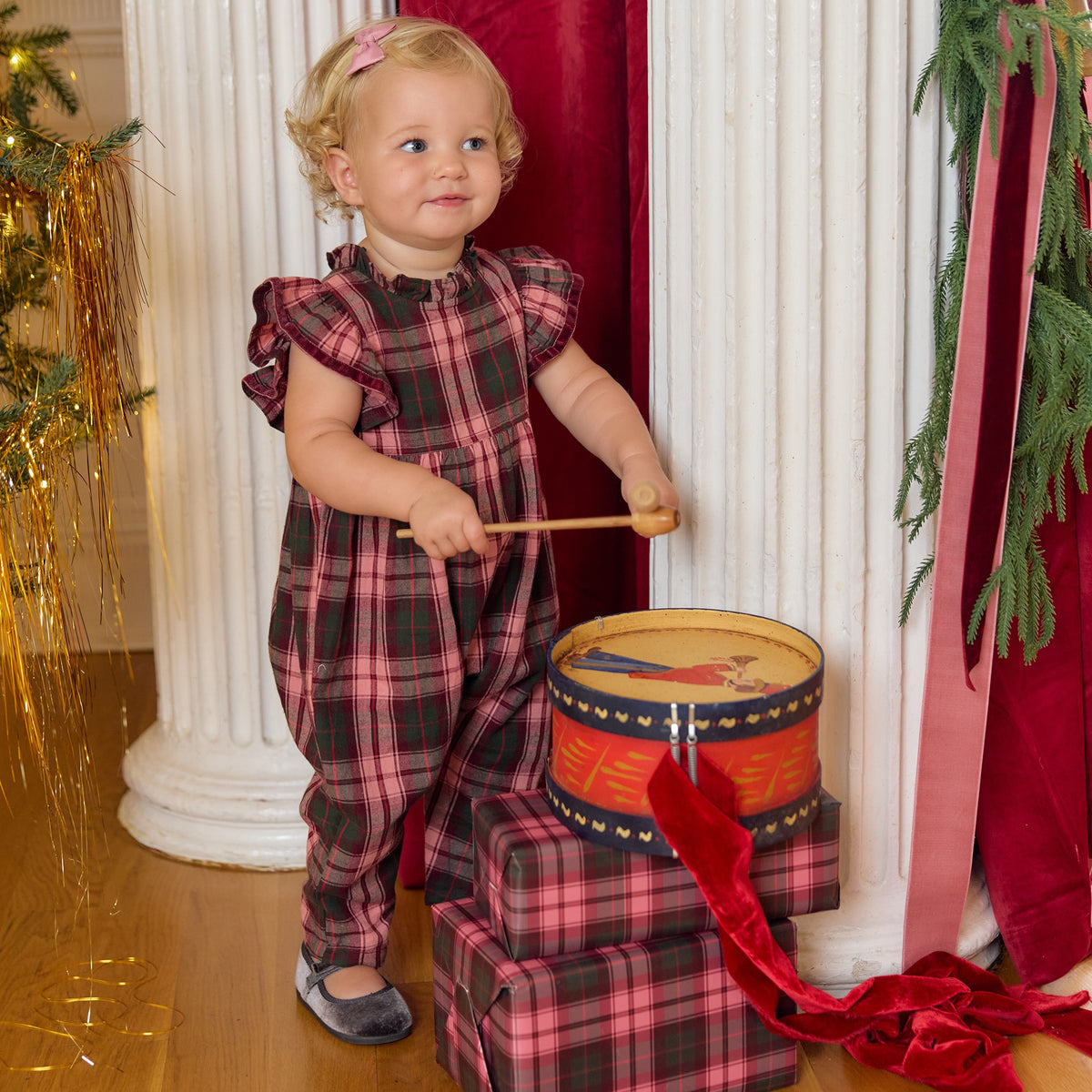 A toddler wearing a Pink Chicken Baby Girls’ Jennifer Jumper in burgundy tartan cotton brushed flannel with ruffles plays a toy drum.