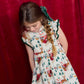 A girl with a side braid wears a Pink Chicken Girls’ Jennifer dress featuring a white ruffled design with a festive Christmas print of reindeer and Santa Claus.