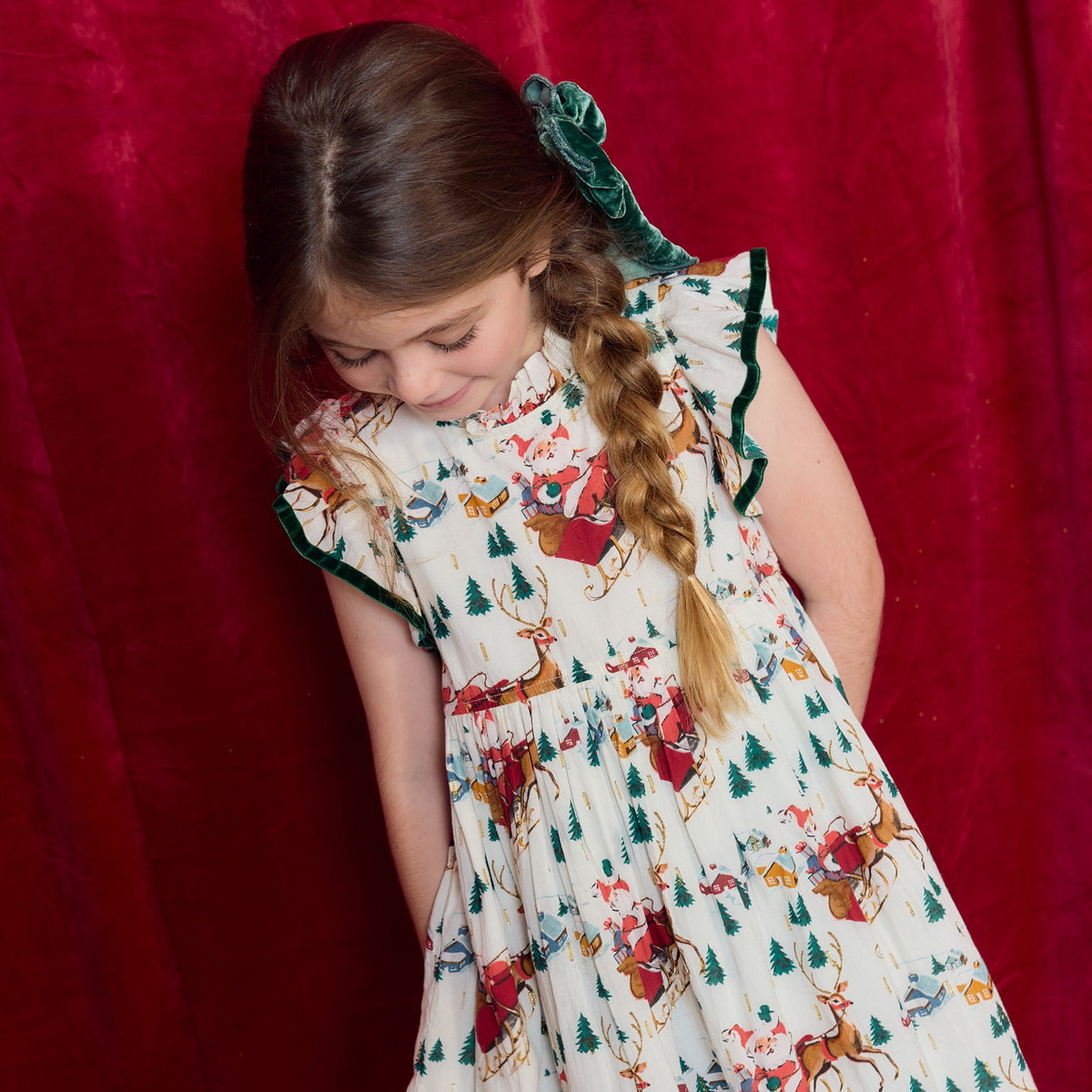A girl with a side braid wears a Pink Chicken Girls’ Jennifer dress featuring a white ruffled design with a festive Christmas print of reindeer and Santa Claus.