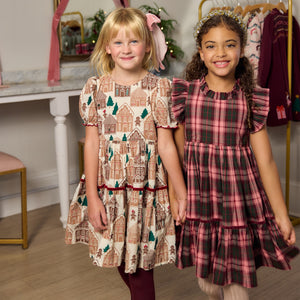 Two girls hold hands, one wearing a tan gingerbread house Eliza Dress and the other in a pink and green plaid tiered holiday dress.