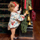 A toddler wearing a Pink Chicken Baby Girls’ Wynne Bubble and a red hair bow touches gold bells hanging from a garland.