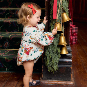 A toddler wearing a Pink Chicken Baby Girls’ Wynne Bubble and a red hair bow touches gold bells hanging from a garland.