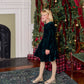 A girl in a Pink Chicken Girls’ Pearl Evergreen Velour dress with ruffles, glittery tights, and gold ballet flats standing in front of a Christmas tree.