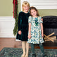 Two young girls standing together, one wearing a dark green velvet Girls’ Hermione Dress and the other in a short-sleeved white Christmas dress with a green holly leaf pattern.