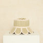 The Manhattan Hat by Lorna Murray, featuring a beige and cream woven design and a scalloped brim, is displayed on a white pedestal against a textured beige background, offering stylish sun protection with UPF 50+.