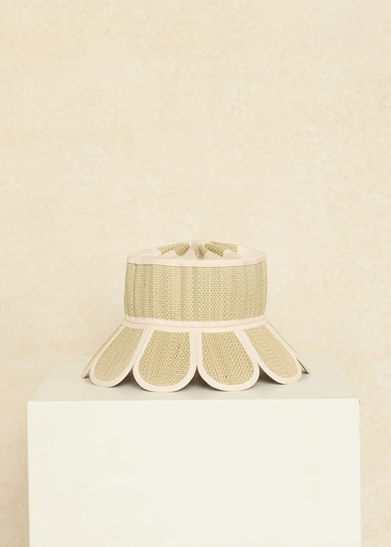 The Manhattan Hat by Lorna Murray, featuring a beige and cream woven design and a scalloped brim, is displayed on a white pedestal against a textured beige background, offering stylish sun protection with UPF 50+.