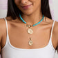 Woman wearing a white top and layered necklaces, including the Jane Win Gumdrop Beaded Necklace Collection with blue beads, a Jane Win pendant, and gold coin accents.