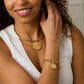 A woman smiles with one hand touching her neck, wearing a white sleeveless top and the Jane Win Destination Pendant Necklace Collection with a matching gold charm bracelet.