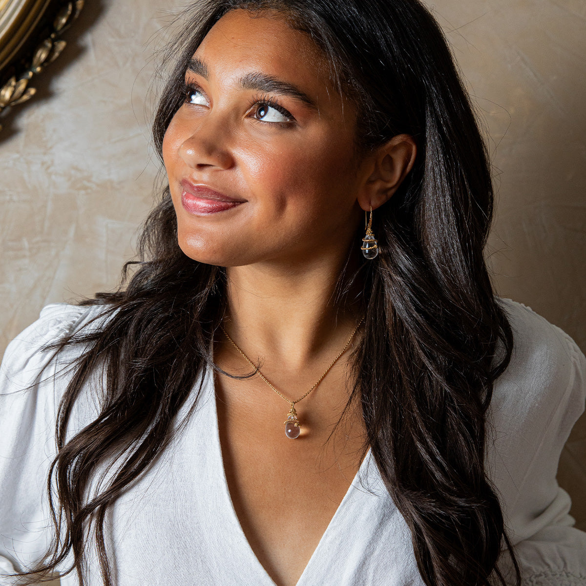 A woman with long dark hair, wearing a white top and Juliska Capucine De Wulf Lily Drop Earrings, smiles softly as she looks up to the left against a textured beige background.