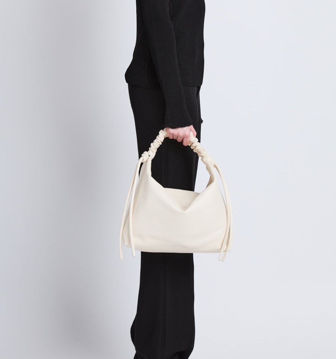 A person wearing a black outfit carries the Proenza Schouler Medium Drawstring Shoulder Bag, which features smooth leather and a twisted handle.