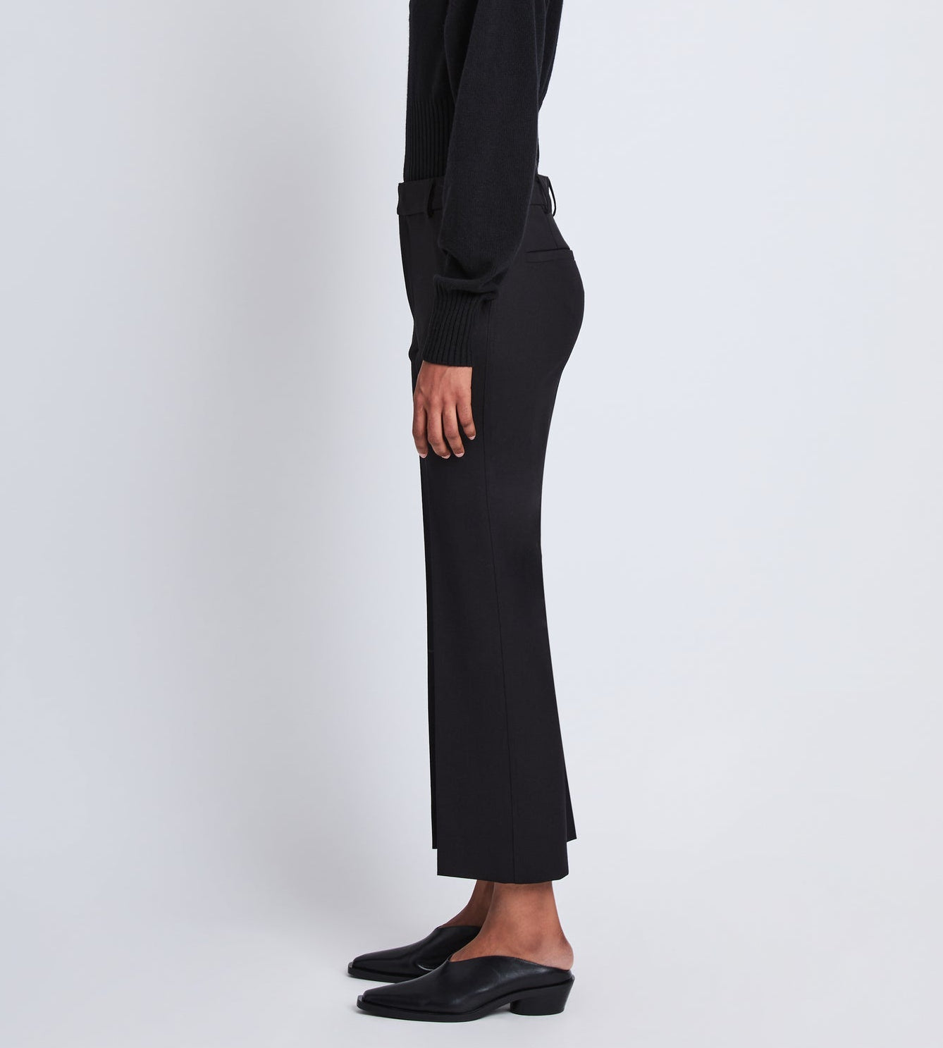 Standing sideways against a plain white background, the person wears the Proenza Schouler Marlene Tropical Wool Pant in black. Their head and upper body are out of frame, and they style the pants with a black cropped sweater and black slip-on shoes.