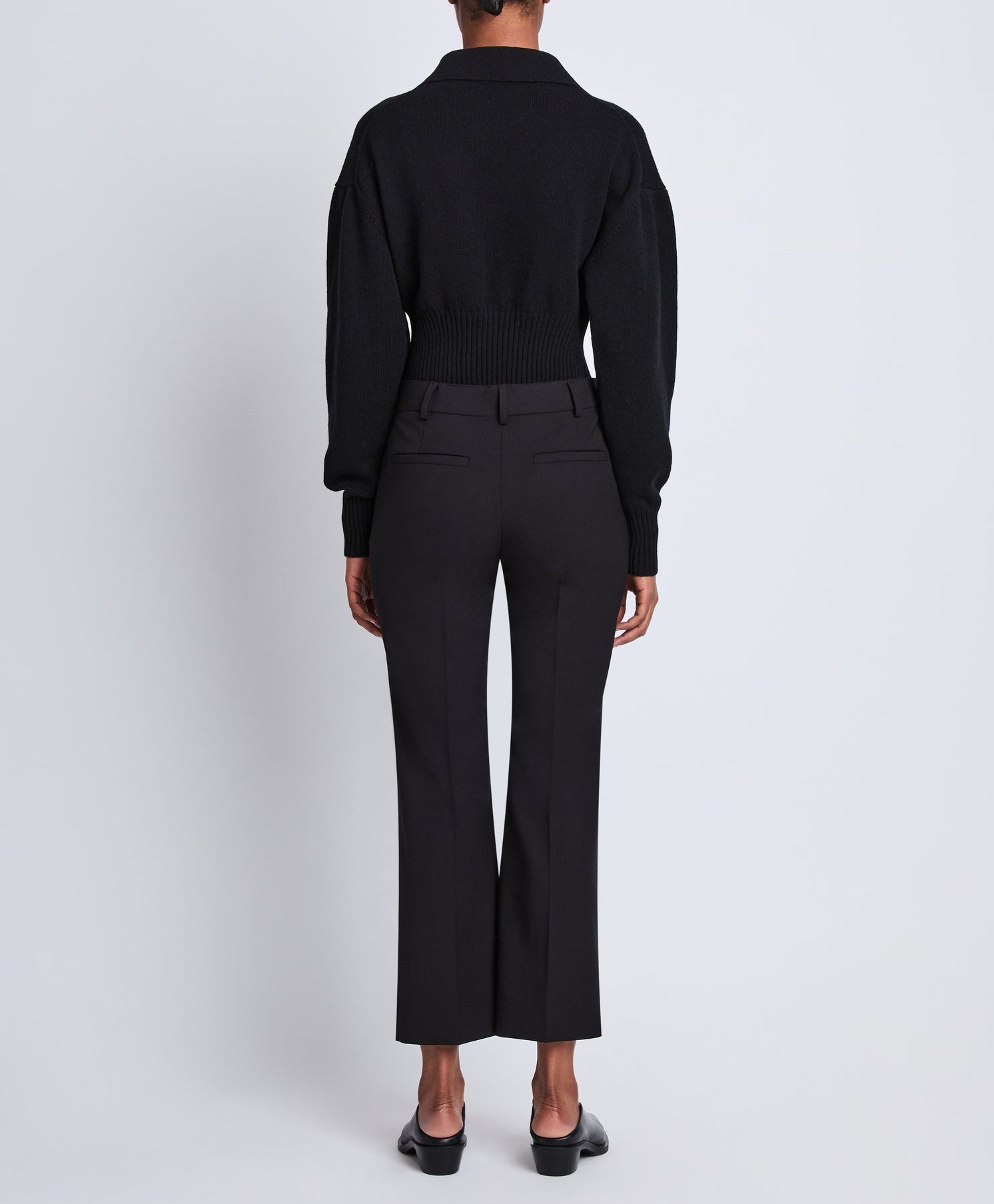 A person stands facing away against a plain white background, wearing a black long-sleeve top, black shoes, and the Proenza Schouler Marlene Tropical Wool Pant—high-rise, tailored, and cropped.